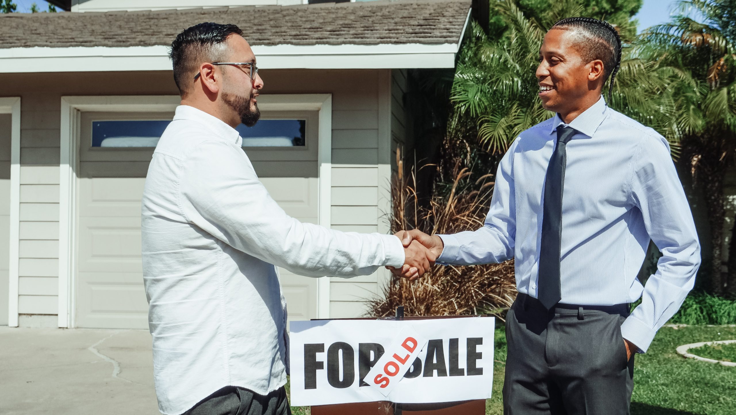 two men shaking hands in front of a house for sale sign